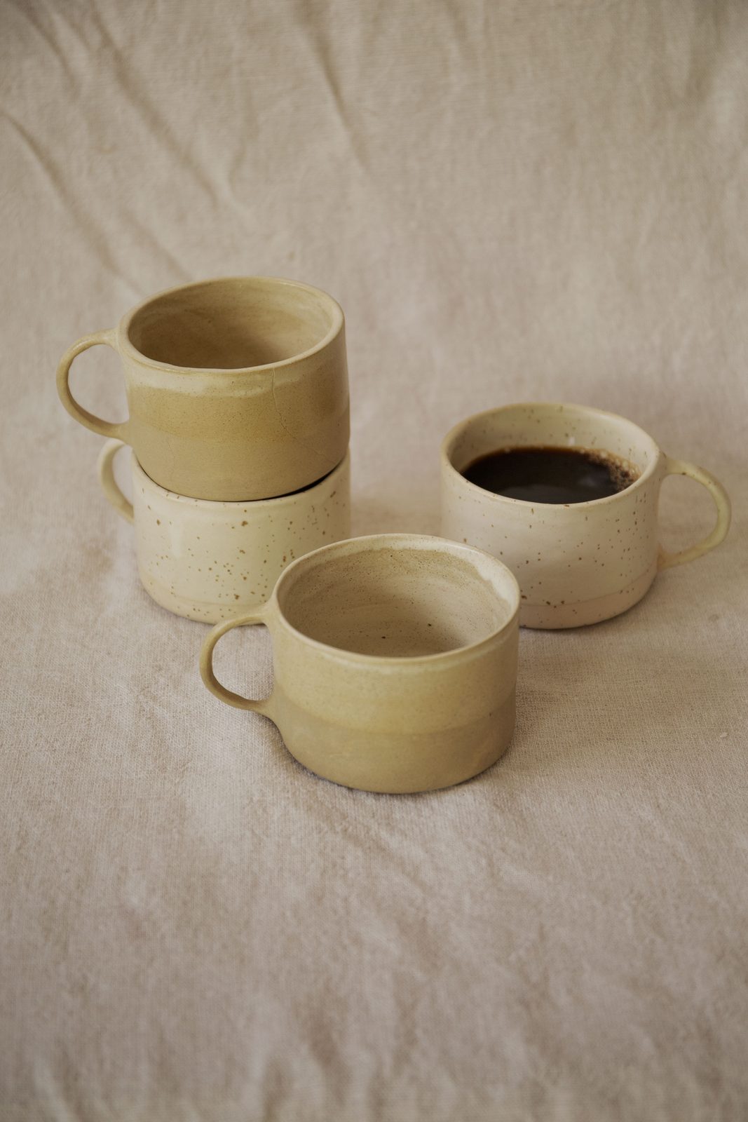 Fyra handgjorda muggar i beige och prickig glasyr på linneduk, en fylld med kaffe
